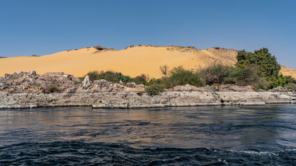 There are picturesque boulders on the river bank. An orange sand dune against a clear azure sky. Waves and foam from a ship on blue water. Egypt. Nile