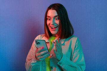 Portrait of amazed surprised Caucasian woman wearing stylish jacket posing isolated on neon light background, using cell phone, reading breaking news, looking at display with big eyes.