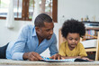 Happy African American family spending time together at home. Cute black son and his handsome father are reading a book while lying on floor in living room. Children education, development concept.