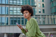 Half Length Shot Of Pensive Woman With Dark Curly Hair Poses At Urbanity With Mobile Phone Downloads New Application For Editing Media Files Checks Notification Dressed In Casual Green Jumper