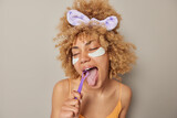 Fototapeta Łazienka - Indoor shot of curly haired young European woman brushes tongue for oral care applies beauty patches under eyes for skin treatment wears casual t shirt and headband isolated over grey background