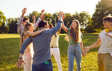 Group Of Happy Young People Have Fun Together While Walking In Park On Warm Summer Evening. Cheerful Excited Multiethnic Friends Laugh Out Loud And Give Each Other High Five.