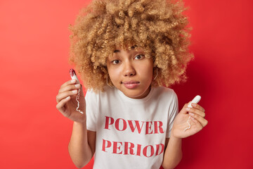 Canvas Print - Menstruation gynecology and hygiene concept. Young curly haired European woman holds two tampons chooses best absorbency product wears white t shirt with inscription isolated over red background