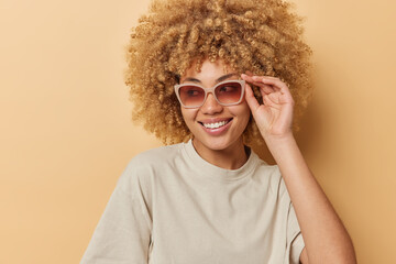 Wall Mural - Indoor shot of happy curly haired young woman wears sunglasses and casual t shirt looks gladfully away has good mood isolated over beige background. Positive human emotions and style concept
