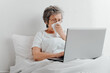 © Sergio - Sick senior woman using laptop while sitting in bed alone in apartment. Elderly woman with runny nose browsing internet or working from home online