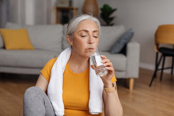 Wall Mural - Domestic sports and hydration. Senior woman drinking fresh water from glass, resting after exercising at home