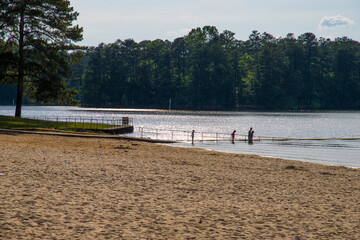 a Mexican family standing in the clear rippling waters of Lake Acworth with silky brown sand, lush green trees, grass and plants with blue sky and clouds at Proctor Landing Park in Acworth Georgia USA