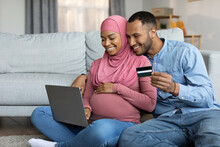 Black Pregnant Muslim Spouses Using Laptop And Credit Card At Home Together