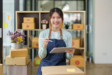 A Portrait Of A Small Startup, An SME Owner, An Asian Female Entrepreneur Checking Orders To Arrange The Produce Before Packing The Products In The Inner Boxes With The Customers. Freelance Concepts