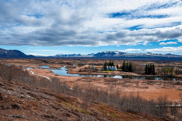 Wall Mural - Distant view of houses amidst stream. Scenic view of beautiful dramatic landscape against cloudy sky. Idyllic picturesque scenery of mountain range and rural land.