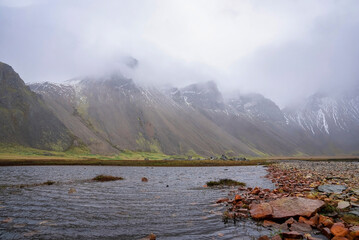 Wall Mural - Close-up of flowing stream amidst field. View of beautiful majestic Vestrahorn mountain peak covered with fog. Idyllic scenery amidst volcanic landscape in valley.