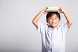 © sorapop - Asian adorable toddler smiling happy wear student thai uniform red pants stand holding book over head and screaming in studio shot isolated on white background, Portrait little children boy preschool