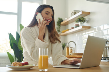 Sticker - Beautiful woman using laptop ant talking on mobile phone while spending time in the kitchen