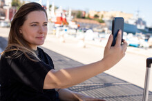 Portrait Of Young Smiling Beauteous Woman Sitting Near Beach Embankment, Taking Selfie In Shadow, Posing On Sunny Day.