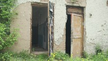 Two Old Wooden Doors With Peeling Paint Swing In Wind Of Abandoned Building On Ruined Stone Wall And Against Background Of Grass And Wood. War, Poverty And Destruction.