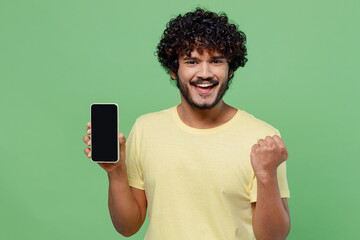 Wall Mural - Young excited cool happy Indian man 20s in basic yellow t-shirt hold in hand use mobile cell phone with blank screen workspace area do winner gesture isolated on plain pastel light green background.