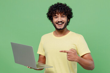 Wall Mural - Young fun happy Indian man 20s in basic yellow t-shirt hold use work point index finger on laptop pc computer isolated on plain pastel light green background studio portrait. People lifestyle concept.