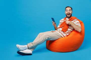 Wall Mural - Full body young smiling fun happy man 20s in orange striped t-shirt sit in bag chair hold point index finger on use mobile cell phone isolated on plain blue background studio People lifestyle concept