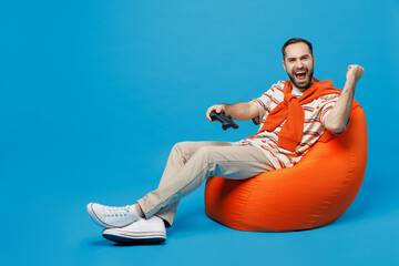 Wall Mural - Full size young excited overjoyed man 20s in orange striped t-shirt sit in bag chair hold in hand play pc game with joystick console do winner gesture isolated on plain blue background studio portrait