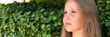portrait face of candid happy little kid girl of eight years old with long blond hair and green eyes on background of green plants during a summer vacation travel. gen z mental health concept. banner