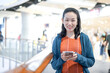 Smiling Asian Female Looks at the camera with holds smartphone in hand, Stands in Department Store Atmosphere.