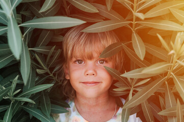 portrait face of candid happy little kid boy of five years old with problem allergic skin and long blond hair and green eyes on background of green plants during a summer vacation travel. flare