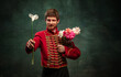 Handsome young man in image of medieval hussar holding flowers isolated on dark blue background. Retro style, comparison of eras concept.