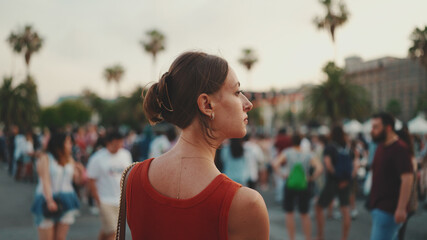 Close-up, girl stands on the square and looks around in the downtown. Closeup of young woman is standing in the city center looking around at the crowd of people in the evening.