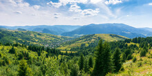Mountainous Countryside Landscape In Summer. Forested Hill And Grassy Meadows On A Warm Sunny Day. Village In The Distant Valley Beneath A Sky With Fluffy Clouds. Transcarpathian Rural Area