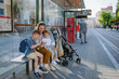 © Halfpoint - Young mother with little kids waiting on bus stop in city, scrolling on mobile phone.