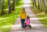 Fototapeta  - Young adult mother and little daughter pushing white baby stroller and walking at town park in warm sunny summer day. Spending time together and breathing fresh air. Enjoying stroll. Two child mom.