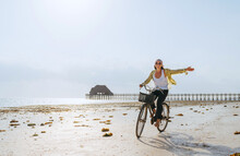 A Young Female Dressed In Light Summer Clothes Joyfully Threw Up Her Hand While Riding Old Vintage Bicycle With A Basket On The Low Tide Ocean White Sand Coast On Kiwengwa Beach On Zanzibar, Tansania
