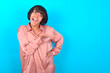 young brunette woman wearing pink silk shirt over blue background says: wow how exciting it is, has amazed expression, shows something on blank space with palm. Advertisement concept.