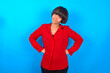 Funny frustrated young brunette woman with short hair wearing red shirt over blue background holding hands on waist and silly looking at awkward situation.