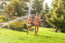 Children Having Fun, Play With Water On Hot Summer Day. Children With Garden Sprinkler. Outdoor Fun. Kids Run Under Water Drops.
