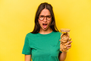 Wall Mural - Young hispanic woman holding a cookies jar isolated on yellow background screaming very angry and aggressive.