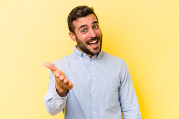 Wall Mural - Young caucasian man isolated on yellow background stretching hand at camera in greeting gesture.