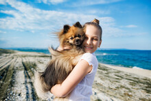 Girl With Blond Hair With Smile Hugs Pomeranian Dog With Golden Hair On Seashore Near Black Sea