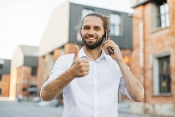 Wall Mural - Portrait of smiling young man doing a phone call showing thumb up. Handsome sporty bearded dark-haired man is using the smart phone outdoor.