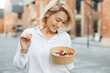 Portrait of a young attractive woman eating salad at the background of buildings of city. Beautiful young female enjoying a fresh salad outdoor. Cute blond girl eating a healthy meal.