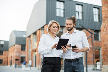 Wall Mural - Charming woman with digital tablet and attractive man with smartphone and cuo of take away coffee standing together near office building. Two colleagues using modern gadgets for work outdoors.