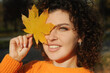 Young smiling woman with autumn leaf has fun in the autumn park. Portrait of curly hair happy woman on the foliage outdoors.