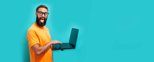 Young crazy bearded charismatic man. Shocked or surprised expression. Laptop concept. Funny promotion poster. Programmer, web developer holding a laptop in his hands and looking at the camera
