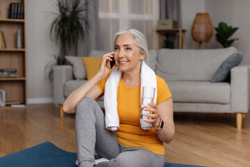 Wall Mural - Happy senior lady with glass of water speaking on smartphone on break from domestic sports training, sitting on yoga mat