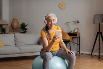 Wall Mural - Happy senior woman with cellphone and wireless headphones exercising on fitball in living room at home, copy space