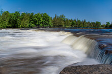 The Blurred Water Of Crowe's Rapids Near Campbellford, Ontario Flows Over Some Rocks On A Bright Sunny Day.