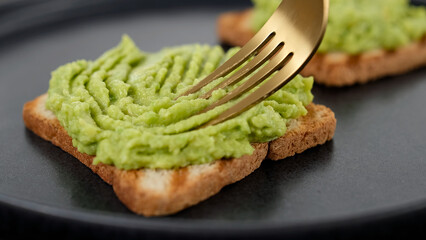 Wall Mural - Preparing avocado toast. Hands with fork spreading mashed avocado on toasted whole  bread. Vegan food concept