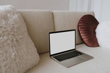 Laptop computer with blank screen on comfortable woolen sofa with pillows. Aesthetic template with mockup copy space. Online store, blog, social media, website branding. Online shopping concept