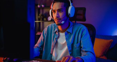 Happy asia guy gamer wear headphone participation play video game colorful neon lights computer in living room at night modern house. Esport streaming game online, Home quarantine activity concept.