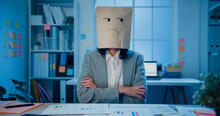 Young Asian Businesswoman Sit On Desk Wear Draw Sad Mask Paper Craft Bag Feel Bad Mood With Fail Work Project In Office At Night. Anonymous Lady Mad And Confuse With Overwork, Work Mental Health.